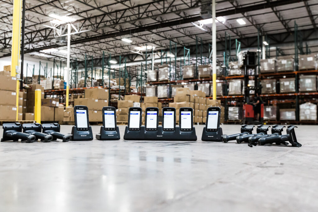 Row of handheld barcode scanners on a warehouse floor with racked inventory behind them